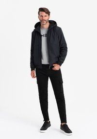 Svart jacka med huva, grå skjorta med svart text, och svarta cargobyxor. Svarta sneakers fulländar outfiten.