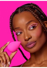 Sponge per il trucco rosa tenuto da una persona con capelli intrecciati. Texture liscia, forma a goccia. Sfondo rosa brillante. Il trucco degli occhi presenta accenti rosa.