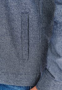 Zbliżenie niebieskiego, teksturowanego rękawa kurtki oraz bocznej kieszeni z delikatnym pionowym rozcięciem, noszonej na niebieskich dżinsach.