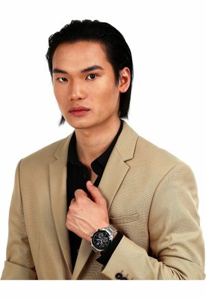 Herreur med sort urskive, sølvfarvet metalrem, båret på en hånd. Personen i en lys beige blazer over en sort skjorte.