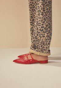 Mules en cuir rouge avec des orteils pointus et plusieurs sangles croisées, associés à un pantalon beige à motif léopard noir.