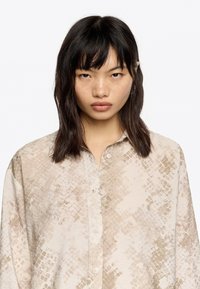 Camisa de manga larga y botones en un tono beige claro con un diseño texturizado de patrón de escamas. Tejido suave con un corte holgado y cuello clásico.