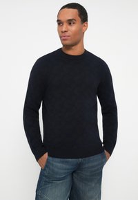 Hombre con un suéter texturizado de color azul marino oscuro y jeans de mezclilla azul, con las manos en los bolsillos, mirando ligeramente hacia la derecha contra un fondo blanco.