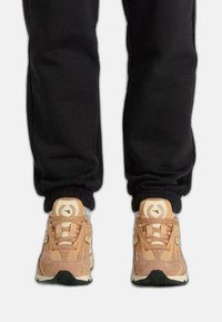 Sneakers gemaakt van beige suede en mesh met een textuurpatroon, met een witte zool en logo-detail, gedragen met zwarte joggers.