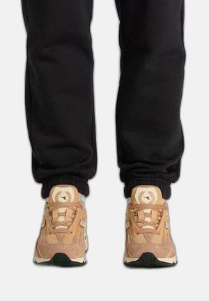Baskets en suède beige et en mesh avec un motif texturé, dotées d'une semelle blanche et d'un détail de logo, portées avec un jogging noir.