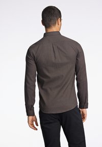 Camicia marrone scuro a maniche lunghe con una trama uniforme, colletto con bottoni e polsini a due bottoni. Progettata con una vestibilità su misura e piega posteriore.