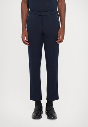 Navy tailored trousers with a slim fit, featuring a button closure, front pleats, and contrasting side detailing. Worn with black loafers.