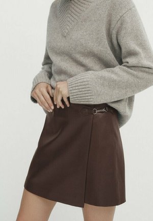 Osoba ubrana w beżowy dziany sweter schowany w brązową skórzaną mini spódnicę z wysokim stanem i bocznym detalem z klamrą.