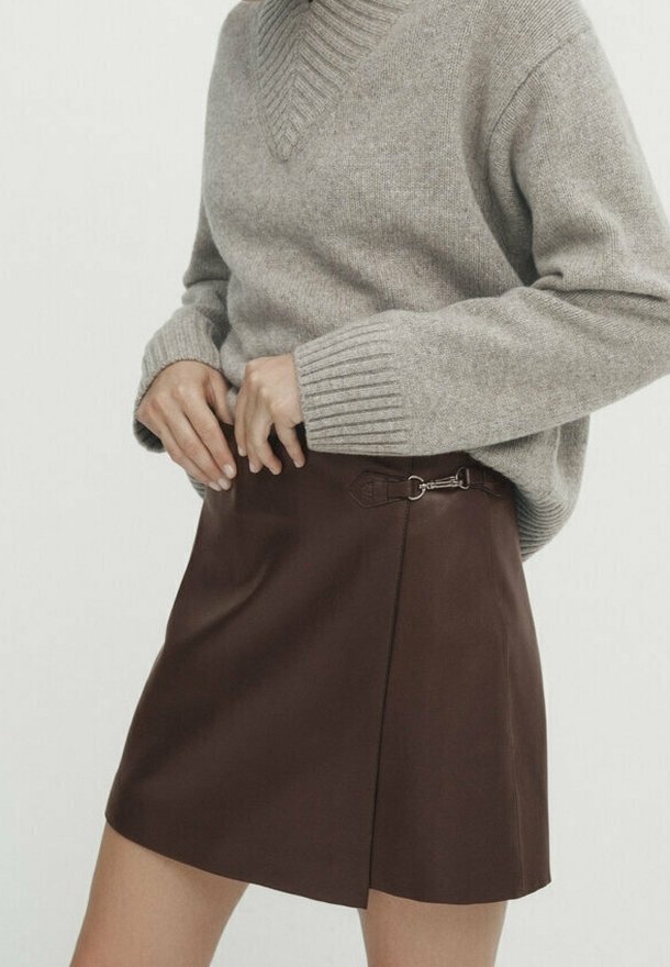 Personne portant un pull en maille beige rentré dans une mini-jupe en cuir marron taille haute avec un détail de boucle latérale.