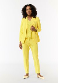 Żółty, dopasowany garnitur z fakturowaną, koronkową górą. Spodnie o wąskim kroju i dopasowana marynarka w połączeniu z beżowymi butami wsuwanymi.