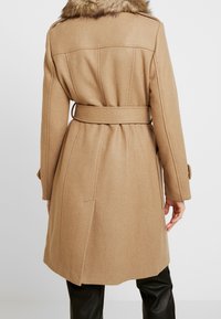 Cappotto di lana cammello con colletto in pelliccia, vita con cintura e vestibilità su misura. Presenta tasche laterali e polsini con bottone. Texture liscia, design strutturato.
