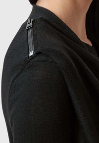 Gros plan d'une personne portant un pull en tricot noir avec un détail de fermeture éclair argentée sur l'épaule, sur un fond uni.