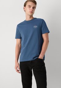 T-shirt in cotone blu con scollo rotondo, caratterizzato da un piccolo logo bianco sul petto. Abbinato a jeans neri scuri.