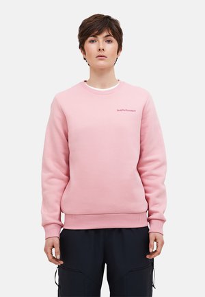 Personne aux cheveux bruns courts portant un sweat-shirt rose clair et un pantalon noir, debout devant un fond clair uni.