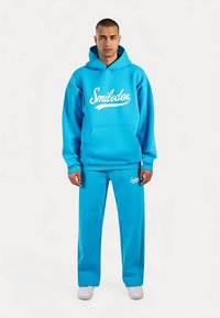 Blauwe hoodie en bijpassende broek van zachte stof. De hoodie heeft een groot wit "Smilodox"-logo en een voorkantzak.