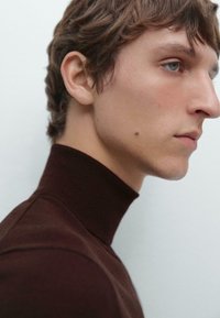 Brun höghalssweater med ribbat mönster som visas på en person. Tyget ser mjukt ut med en jämn textur och har en åtsittande design. Bakgrunden är ljus.