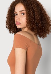 Top castanho sem mangas, com mangas curtas, feito de tecido macio. Apresenta um design ajustado e um pequeno detalhe em laço nas costas.