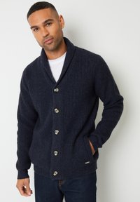 Cardigan in maglia a coste blu navy con collo a scialle, cinque bottoni e tasche laterali; presenta un tessuto strutturato e un dettaglio logo sottile all'orlo.