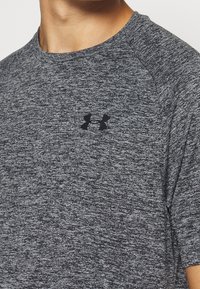 Šedé, texturované sportovní tričko s krátkým rukávem a kulatým výstřihem, se vyznačuje černým logem Under Armour na hrudi.