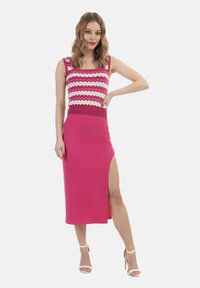 Abito rosa a costine con uno spacco alto, caratterizzato da un top a zigzag rosa e bianco. La modella indossa tacchi con cinturini, in piedi con una mano posata sull'anca.