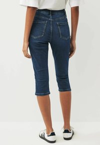 Denim capribyxor i mörkblått, med hög midja, fem fickor och kort längd. Slutet är rullat. Bärs med vita sneakers.