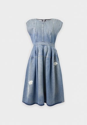Frau trägt ein blaues Denim-Midikleid mit Kappärmeln, Blumenstickerei und Taschen, kombiniert mit weißen spitzen High-Heels.