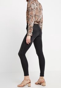 Jean skinny noir en matière extensible, avec taille haute et poches arrière, portés avec des sandales à talons blocs beiges et un haut à imprimé serpent.