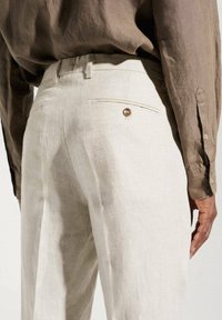 Mann trägt beiger Leinenhose mit einer knöpfbaren Gesäßtasche und einem braunen langen Hemd, das eingesteckt ist.