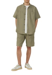 Khaki Kurzarmhemd mit zwei Brusttaschen, Knopfleiste und darunter einem weißen T-Shirt mit Rundhalsausschnitt; kombiniert mit passenden Shorts und Turnschuhen.