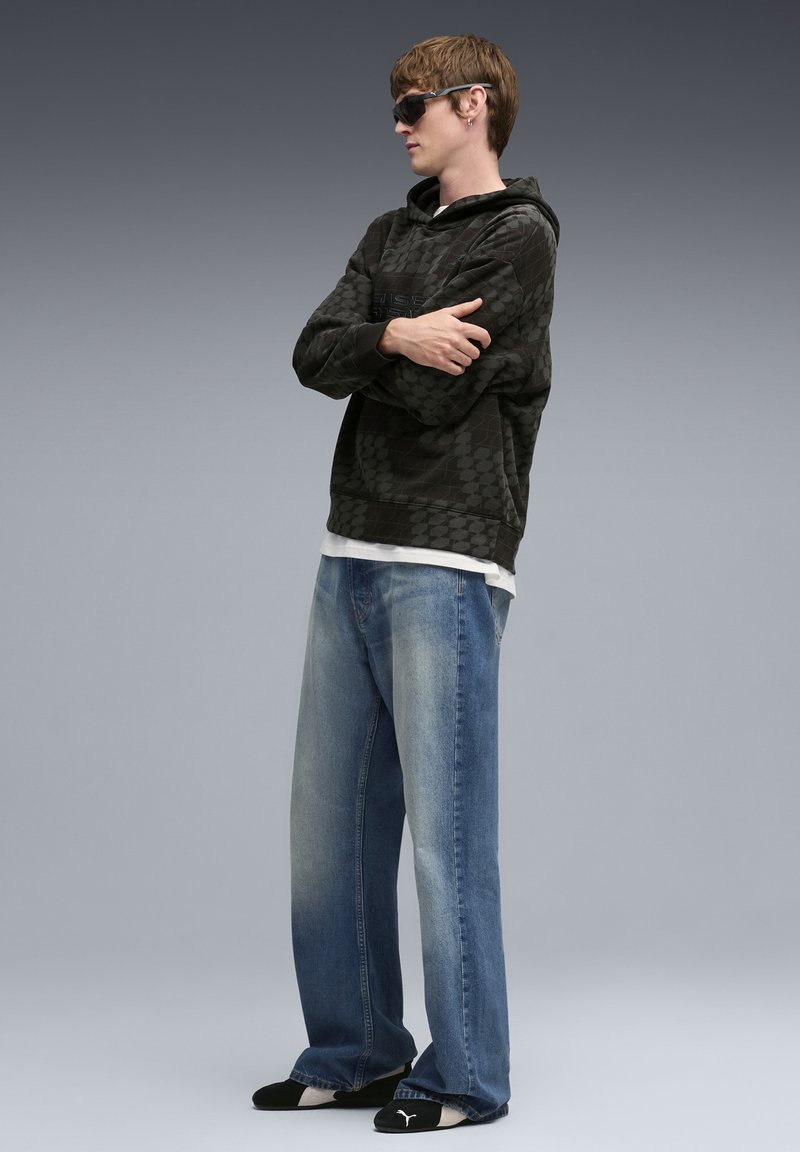 Felpa con cappuccio scura a motivo con coulisse, jeans larghi azzurri chiari e scarpe slip-on nere. Il modello indossa occhiali da sole e sta in piedi con le braccia incrociate.