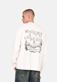 Long-sleeved, off-white t-shirt with black graphic text design on the back, featuring "MULTIPLY APPAREL." Soft cotton fabric, relaxed fit.