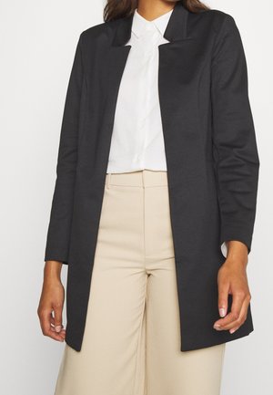 Kobieta ubrana w czarną marynarkę z otwartym przodem, białą koszulę zapinaną na guziki i beżowe spodnie z wysokim stanem na jednolitym tle.