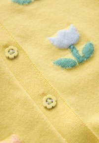 Dzeltens adīts kardigans ar ziedu rakstu, kurā ir zilas un zaļas krāsas akcents. Priekšpusē ir dzelteni ziedu formas pogas.