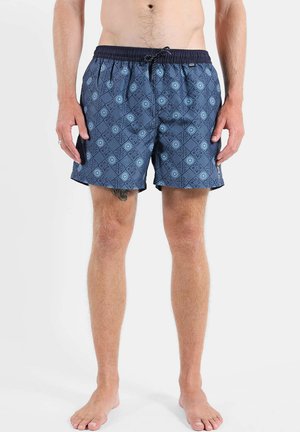 Homme portant un short de bain bleu à motifs avec un cordon noir, debout pieds nus sur un fond blanc uni.