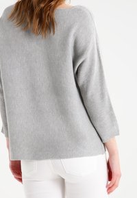 Grå ribbad stickad tröja med vid halsringning och trekvartsärmar, kombinerad med vita byxor. Mjuk textur, avslappnad siluett.