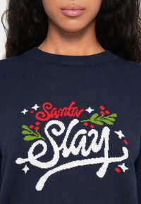 Marinblått stickat tröja med en festlig design där det står "Jultomten Slede" i röd och vit med gröna hollyblad och bär som accent.