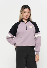 Fialová skrátená mikina s čiernymi raglánovými rukávmi, štvrťový zips, biele prúžky a zlatú nášivku s logom "ROXY" na hrudi.