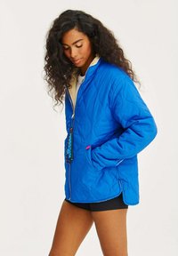 Prošívaná modrá bunda s béžovým interiérem, zipovým zapínáním a dvěma bočními kapsami. Mezi akcentní detaily patří barevná táhla na zip a boční rozparky.