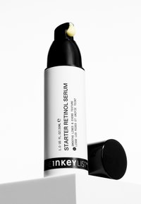 Flacon pompe blanc et noir de sérum rétinol Starter avec crème sur le bec, bouchon noir à côté, posé sur une surface blanche sur fond blanc.