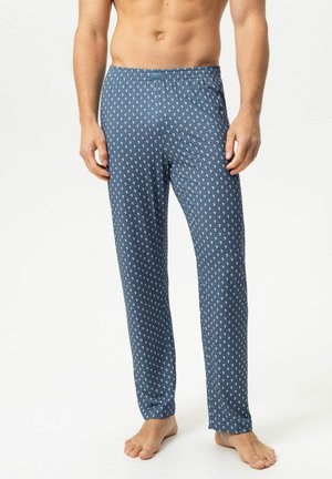 Mann in blau gemusterten Pyjamahosen steht barfuß vor einem schlichten weißen Hintergrund.