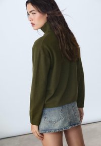 Giovane donna con lunghi capelli scuri che indossa un maglione a collo alto verde oliva e una mini gonna di jeans sbiadita, rivolta a sinistra.
