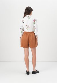 Korte bruine suède shorts met een elastische tailleband, gecombineerd met een witte blouse met bloemenprint en uitlopende mouwen.