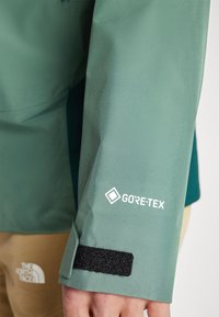 Veste GORE-TEX verte avec une finition texturée, dotée d'une bande Velcro au niveau du poignet et d'un logo imprimé en blanc.