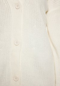 Cardigan tricotat crema cu un model texturat, guler în V și cinci închideri cu nasturi din plastic neted. Material moale și ușor.