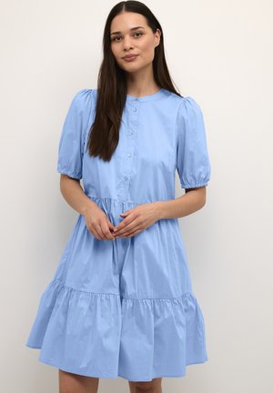 Woman with long dark hair wearing a light blue button-up dress with puffed short sleeves and a tiered skirt, standing against a plain background.
