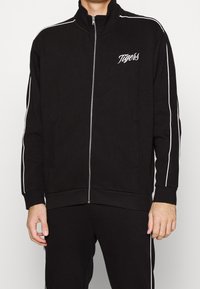 Veste de survêtement noire zippée avec col montant et bandes blanches sur les côtés, avec "Tigers" brodé sur la poitrine et les poches avant.