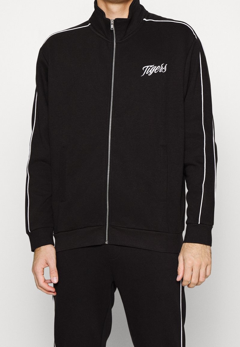 Veste de survêtement noire zippée avec col montant et bandes blanches sur les côtés, avec "Tigers" brodé sur la poitrine et les poches avant.