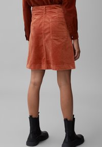 Billie-jupe en orange rouille, dotée d'une poche arrière, de forme évasée et en tissu texturé. Portée avec des bottines noires.