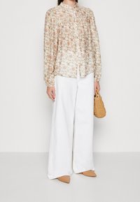 Kvinde iført en lys blomsterbluse, hvide brede bukser, beige spidse sko og holder en lille flettet håndtaske.