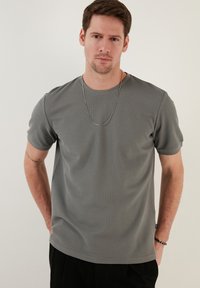 Tricou gri cu mânecă scurtă, dintr-un material neted și texturat. Dispune de un guler rotund și un design minimalist, însoțit de un colier din lanț argintiu.